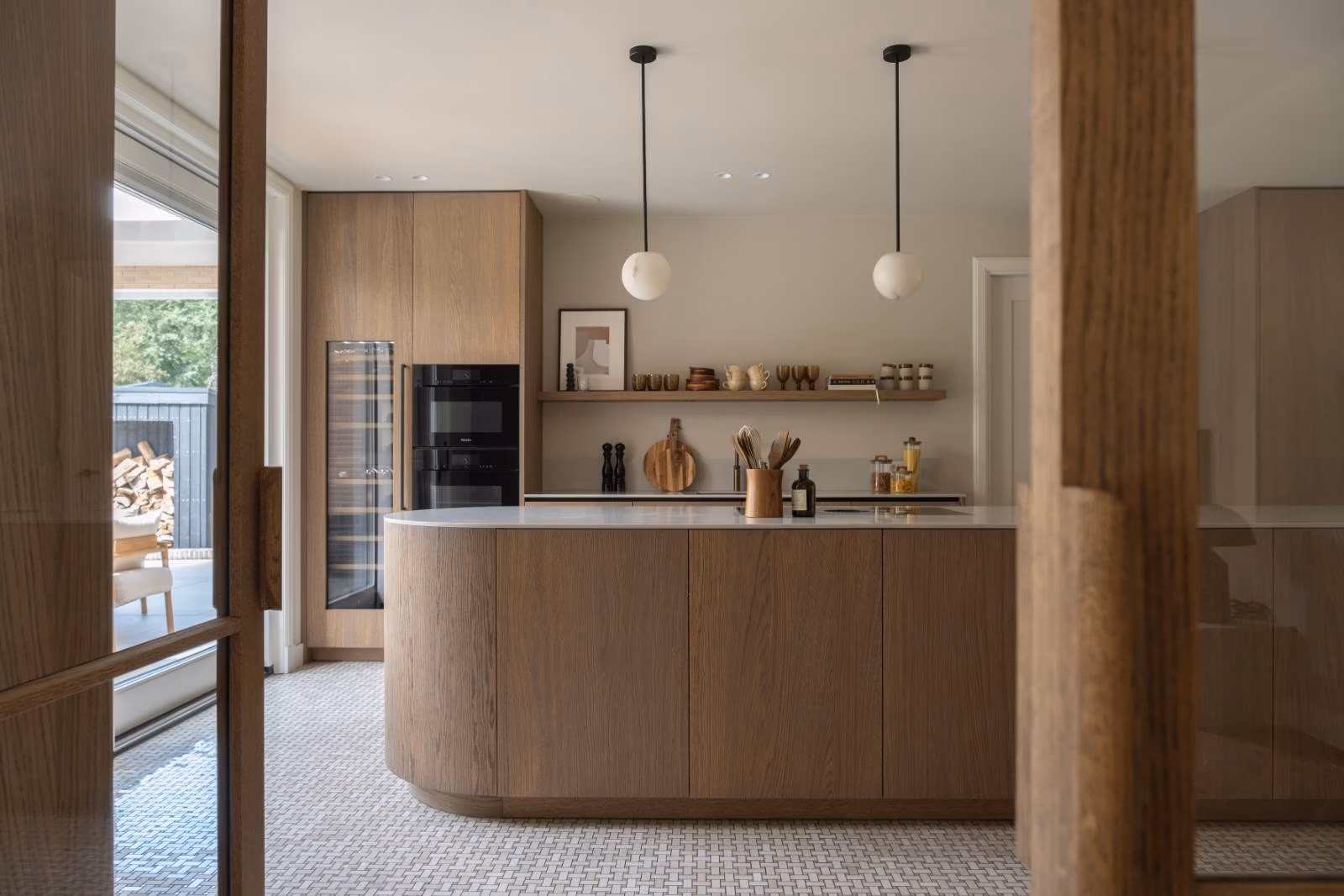 Keuken op maat in Laren in optima forma: elegant rond eiland met wit werkblad, warme houten kastfronten, stijlvolle hanglampen en open schappen met decoratieve accenten; een harmonie van licht, ruimte en verfijning.