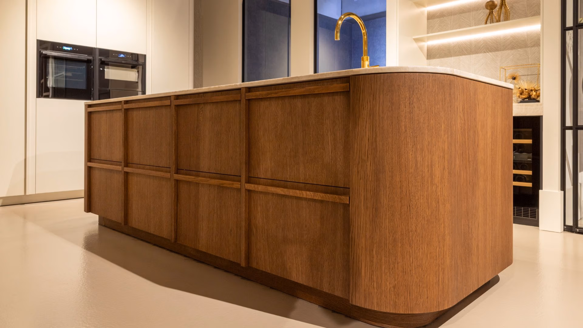 keukeninterieur in de showroom van By Sensa, met een centraal keukeneiland van hout en marmeren blad, goudkleurige kraan, ingebouwde ovens en minimalistische afwerking in warme tinten.