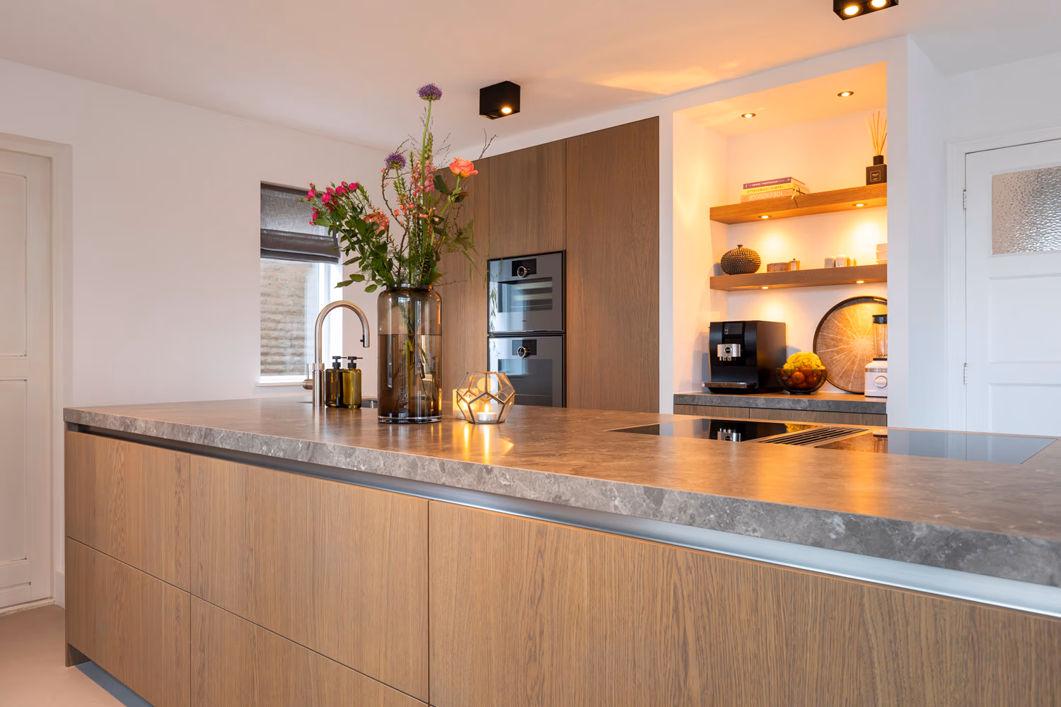 Grijs-bruin eikenhouten kookeiland met keramisch werkblad, spoelbak en hoge kastenwand met Gaggenau ovens.